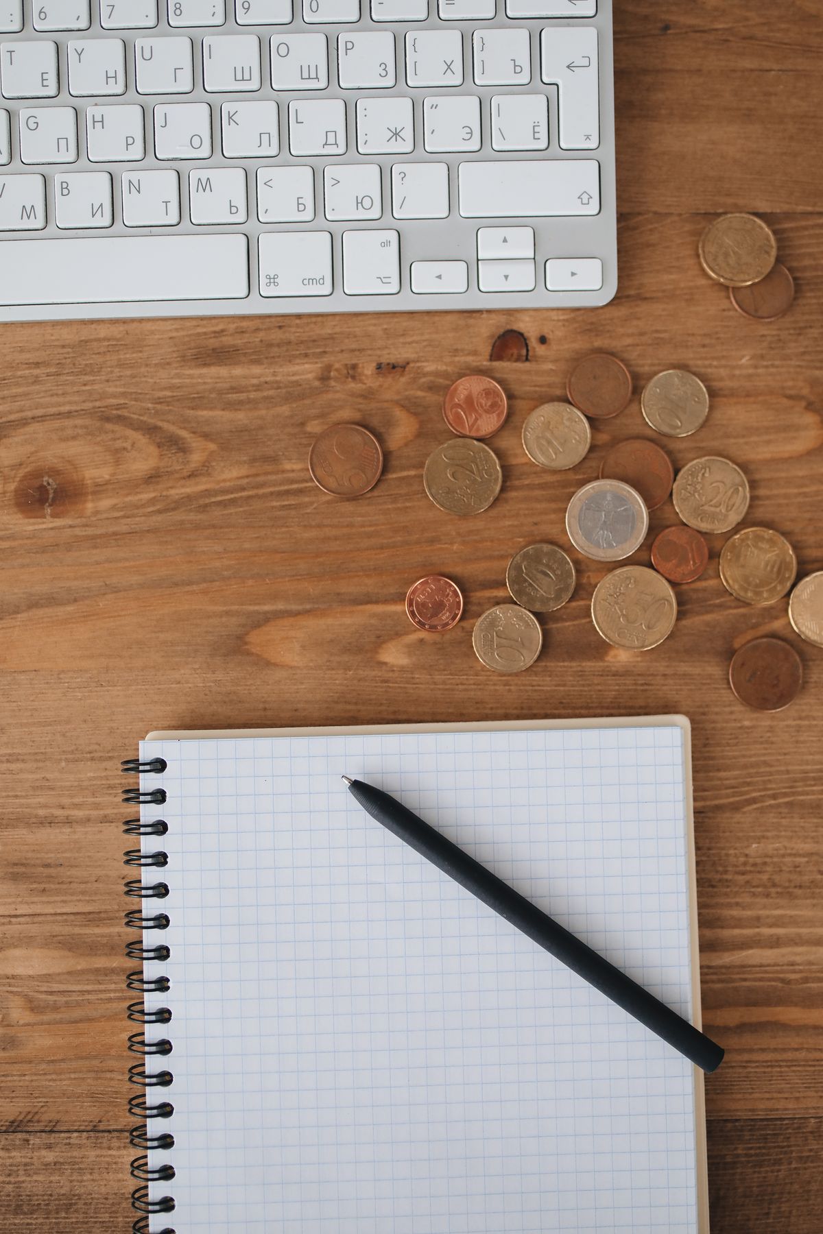 Overhead view of a workspace with keyboard, coins, and a notebook on a wooden desk